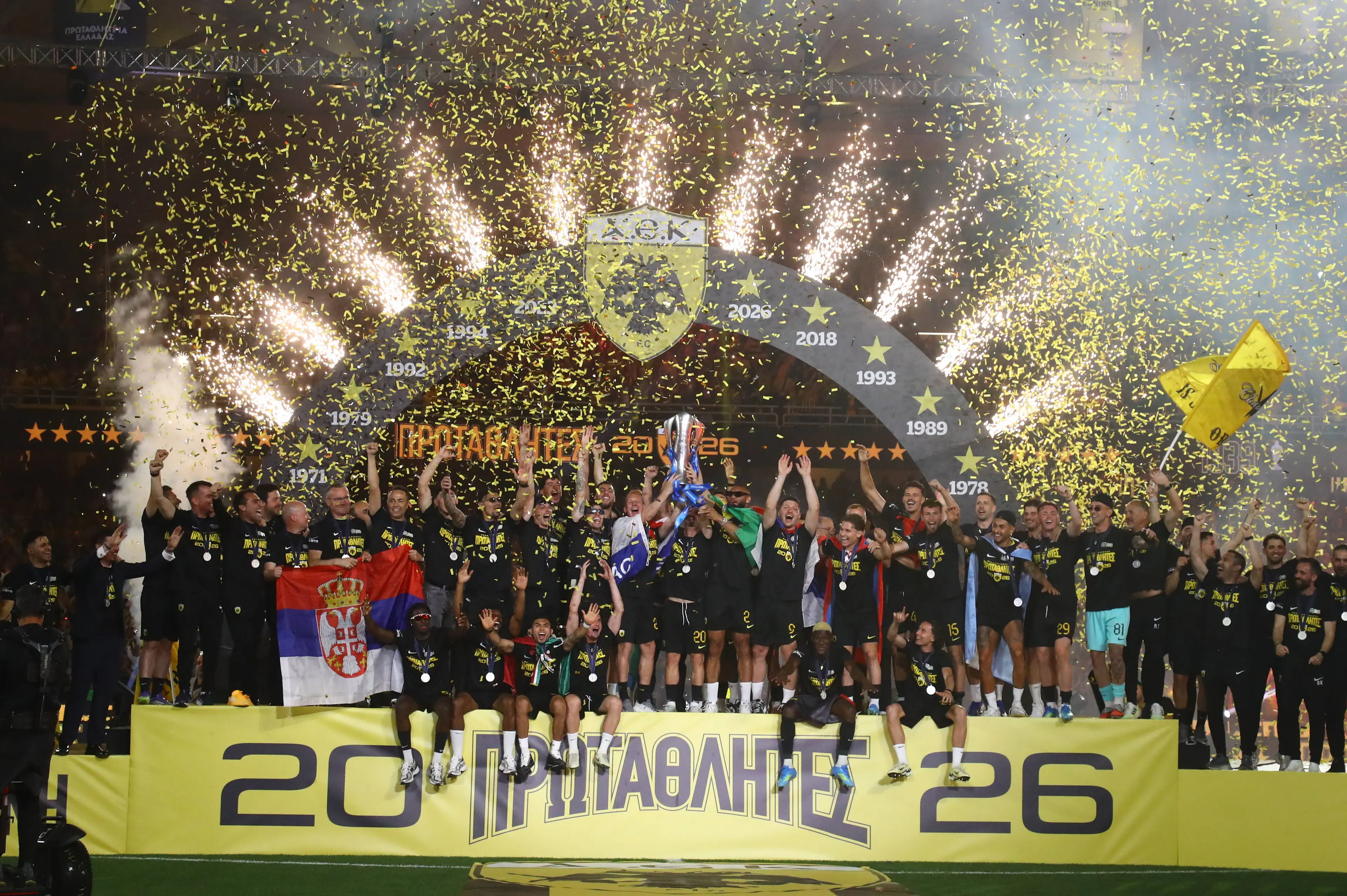 Η AEK πανηγυρίζει την κατάκτηση της Super League 2025-26, σηκώνοντας το τρόπαιο του πρωταθλητή μέσα σε ένα εντυπωσιακό σκηνικό με κίτρινα κομφετί και ξέφρενους πανηγυρισμούς της ομάδας.