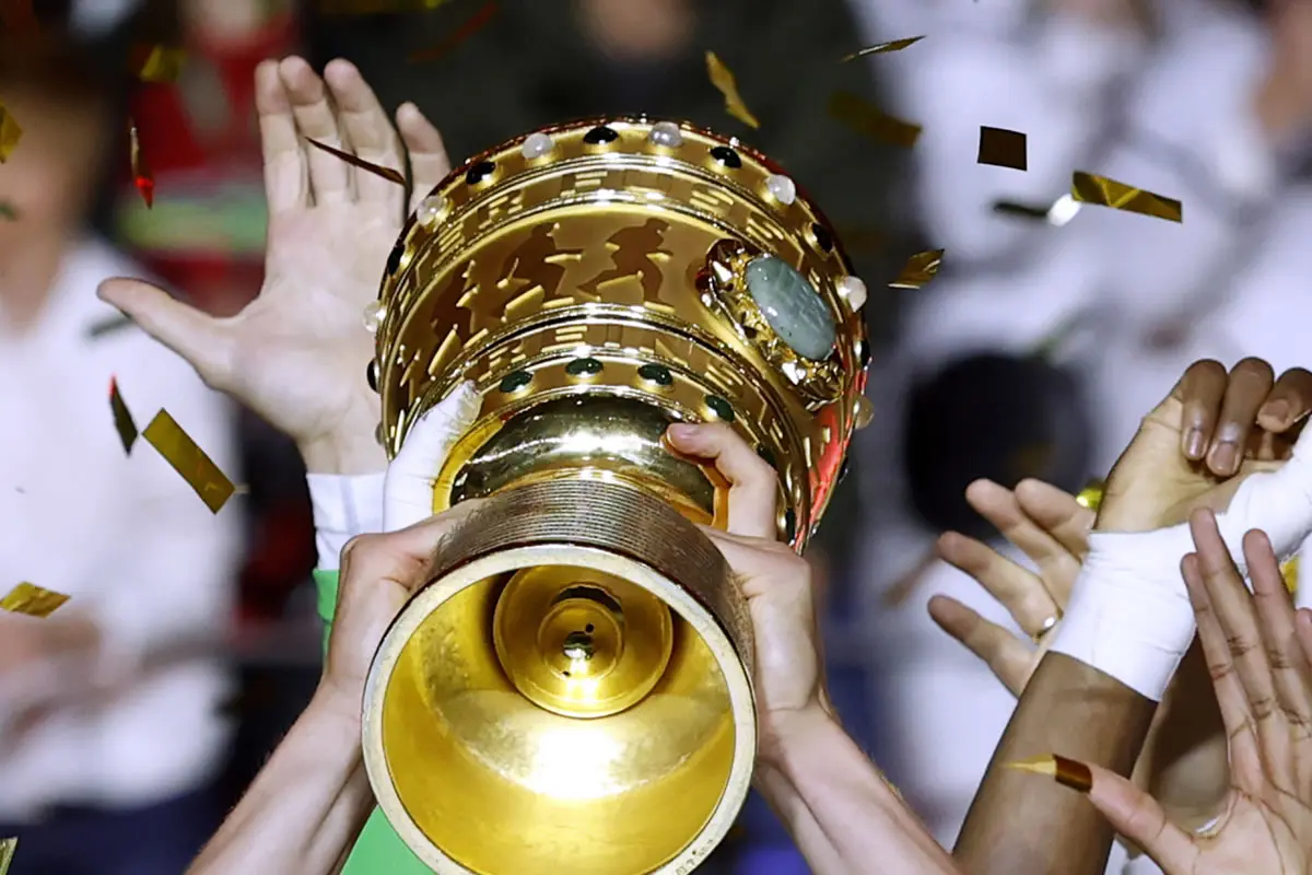 Zwei Hände stemmen den Pokal in die Höhe. Im Hintergrund sind weitere Hände und Konfetti.